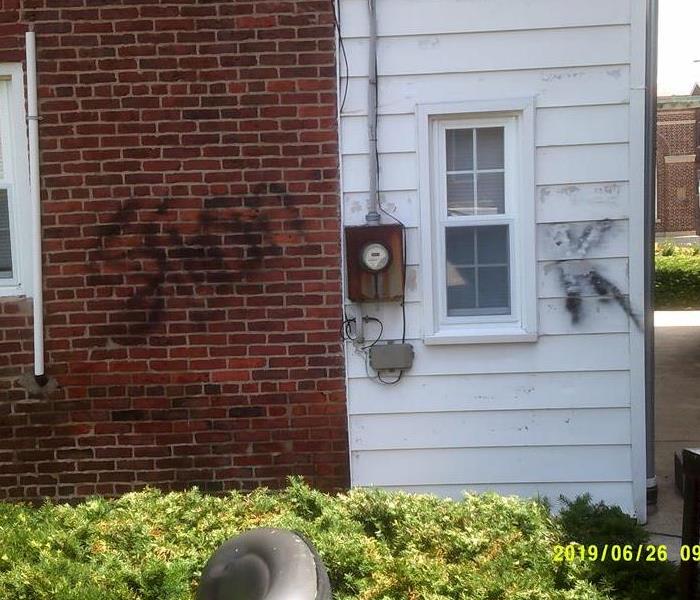 The side of a building has been vadilized with black graffiti on the white siding and brick